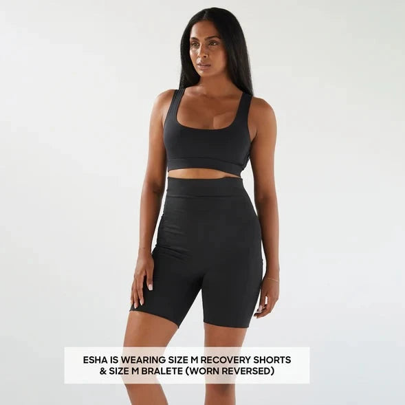 A woman in a black sports bra and Bare Mum Postpartum Recovery Shorts standing against a light background, labeled with her outfit details.