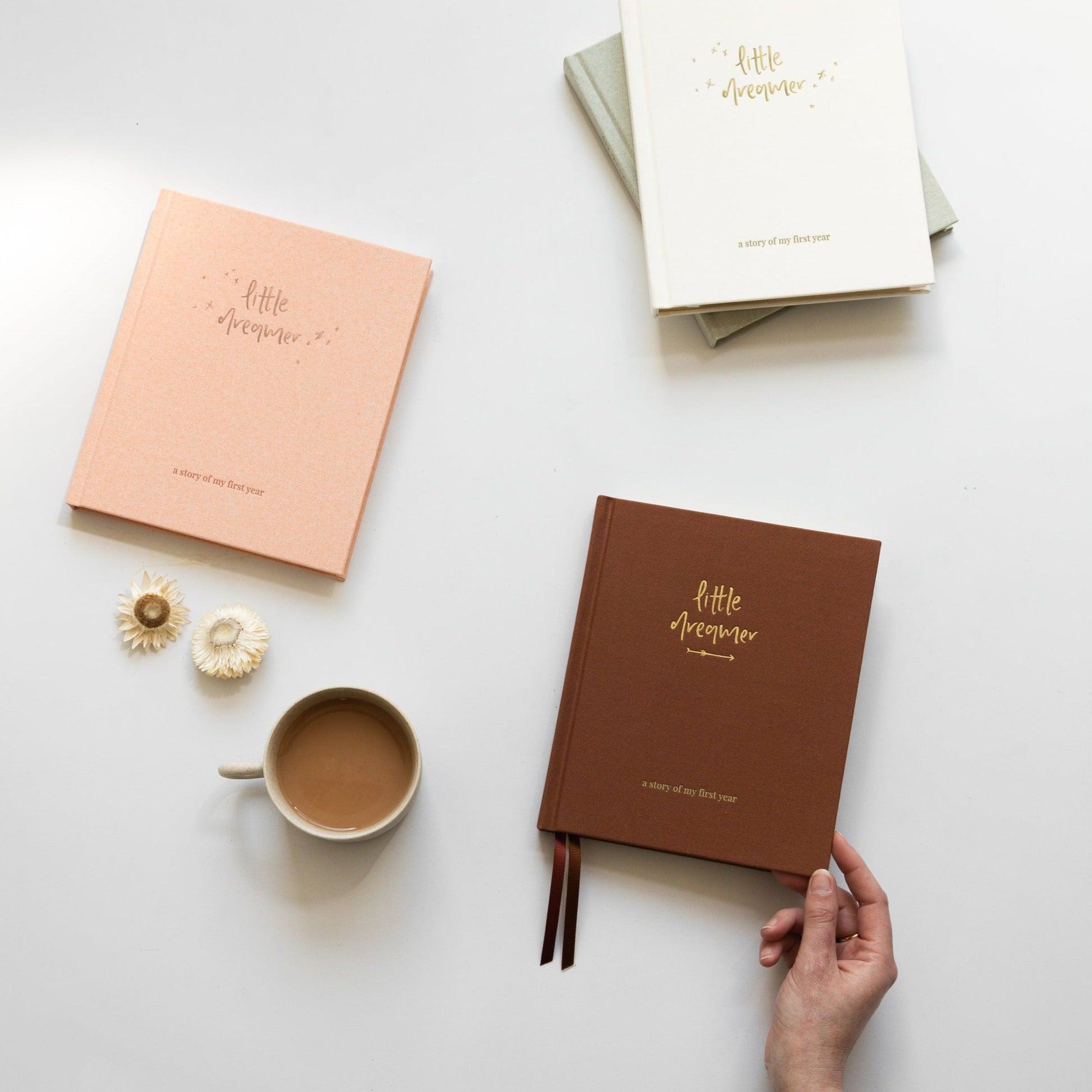 A person holding a Emma Kate Co. Little Dreamer Baby Journal next to a cup of coffee surrounded by other journals.