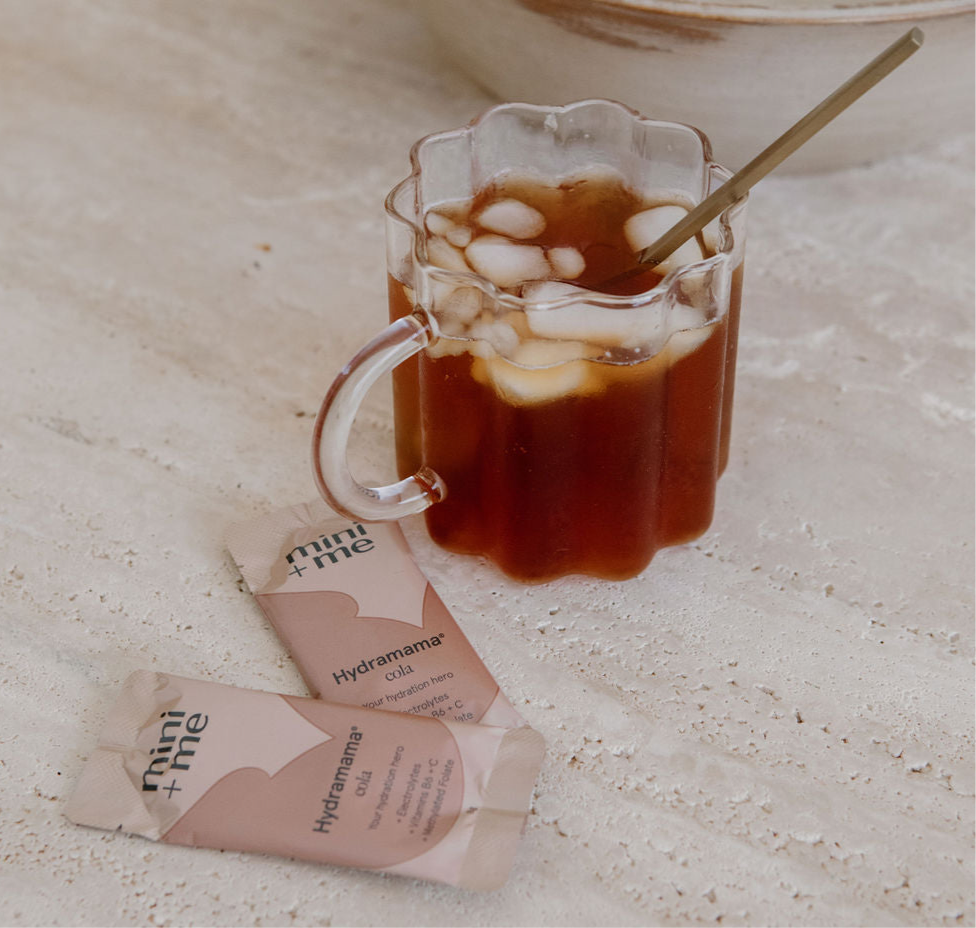 A glass filled with iced coffee and a metallic straw is placed beside two packets labeled "Mini+Me Hydramama Cola" on a light surface, ideal for postpartum hydration with extra electrolytes.