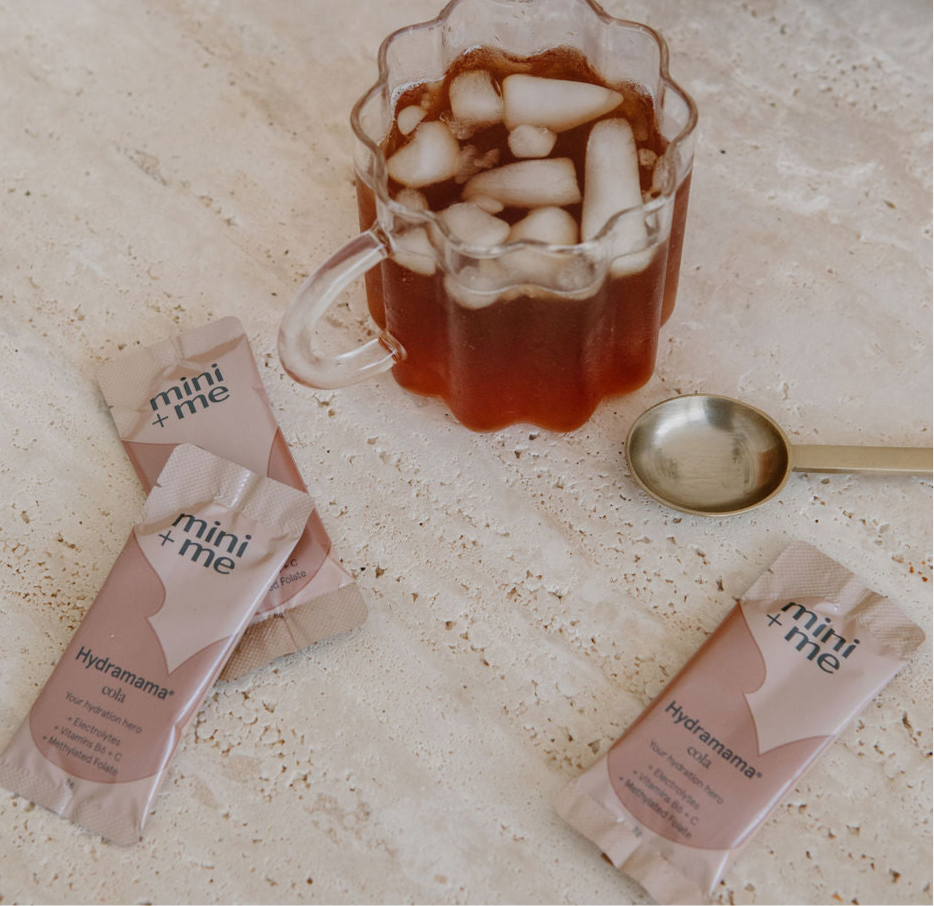 A glass mug filled with iced tea sits beside three pink sachets labeled "Hydramama Cola" from Mini+Me, perfect for a refreshing postpartum hydration beverage, with a small silver spoon resting on the beige countertop.