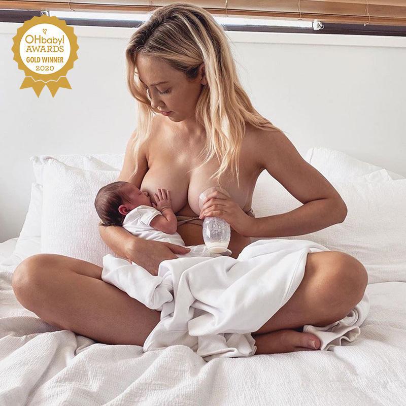 A woman is using the Haakaa Silicone Breast Pump 150ml Gen 2 whilst breastfeeding her baby on a bed.