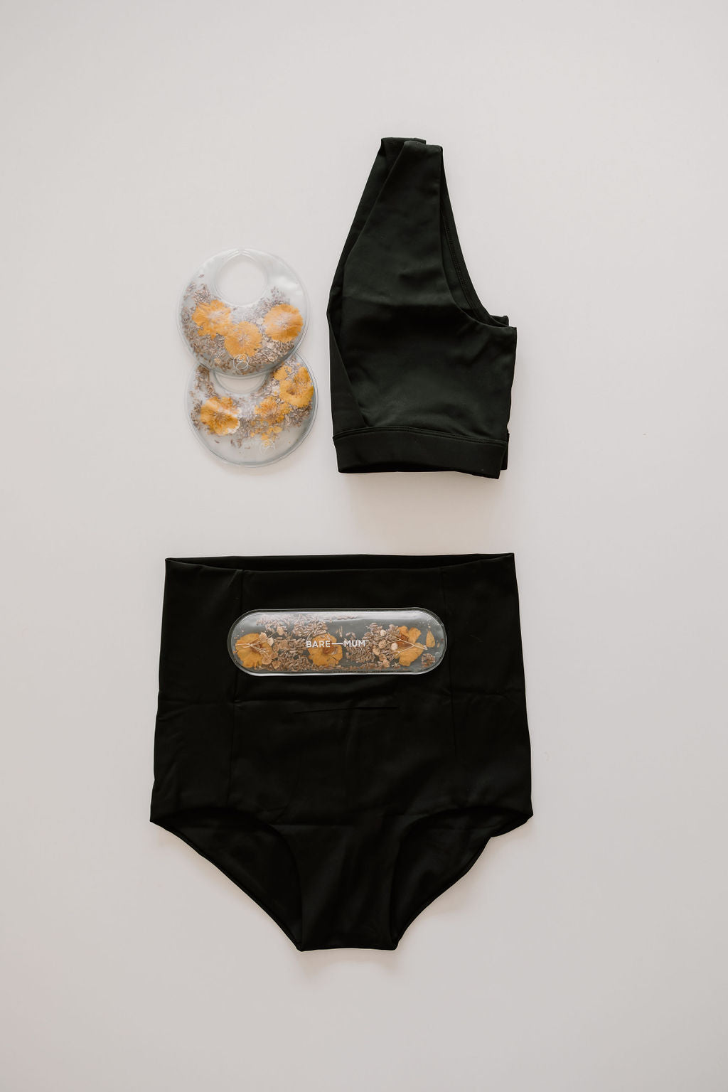 Black sports bra and high-waisted recovery shorts with a small transparent bag of flowers. Two round flower-filled bags are also placed next to the bra. Items, part of a Bare Mum bare mum recovery kit, are arranged on a neutral background.