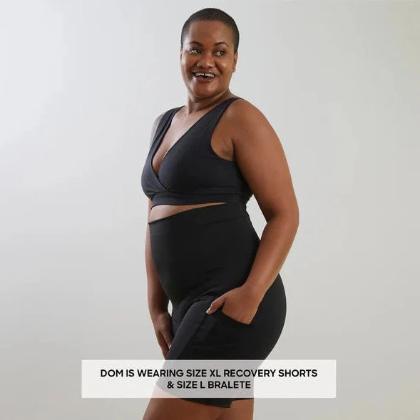 A confident woman in a black bralette and Bare Mum Postpartum Recovery Shorts smiling at the camera, with text indicating her clothing size.