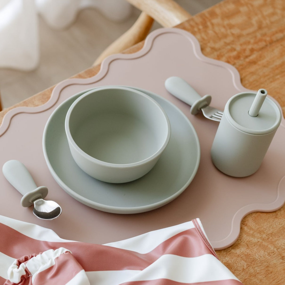 The children's dinnerware set, placed on a pink placemat, showcases premium silicone items like a bowl and plate alongside Rommer's sippy cups from the 2-pack set. A spoon, fork, and striped cloth napkin are in the foreground to ensure a non-toxic mealtime experience.